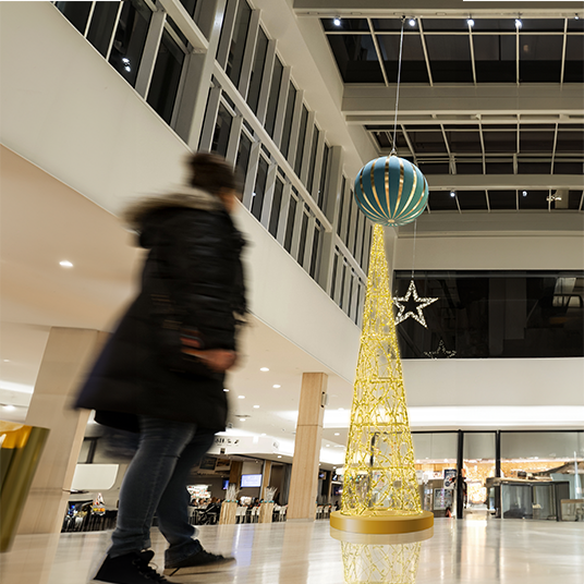 This is a Leavesden Tree inside a mall, which is featuring a cone shape, adorned with gold metal braiding and golden leaves that elegantly wind around the tree. The tree is illuminated with warm white lights, casting a festive glow.