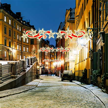 An overhead decorative star swirl on a snowy street attached between 2 European style buildings. With a flowing ribbon design, illuminated in gold and red.