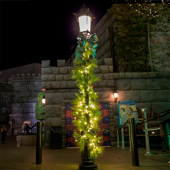 A lush Torrey Pine garland being displayed on a black light pole. Featuring realistic needles in a vibrant green hue. Warm white lights peek through the dense foliage.