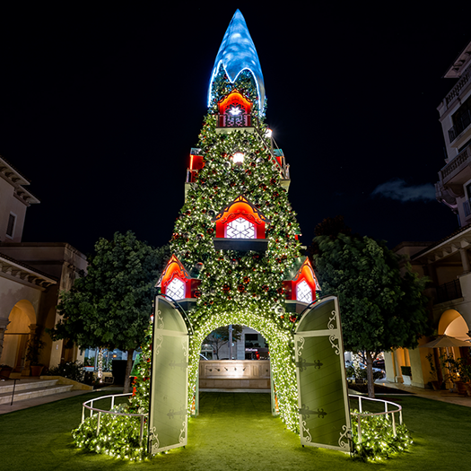 A large 40' Sequoia Tree, with a wide base and narrow top. The tree is decorated with many strands of warm white lights. The center of the tree is hollowed out to create a tunnel-like opening. The tunnel is decorated with lights, creating a winter wonderland scene.